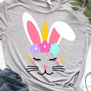 May include: A gray t-shirt with a white bunny rabbit face with pink ears, a yellow unicorn horn, and a flower crown with purple, pink, and blue flowers. The bunny has closed eyes, a pink nose, and black whiskers.