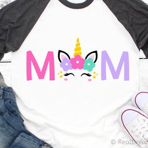 May include: A white t-shirt with a black and grey raglan sleeve design. The shirt features a pink, purple, and blue unicorn with a gold horn and a flower crown. The word "MOM" is written in pink and purple letters.