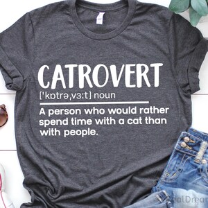 May include: Dark gray t-shirt with the word "CATROVERT" in large white letters. The definition below reads: "A person who would rather spend time with a cat than with people." Sunglasses and jeans are visible.