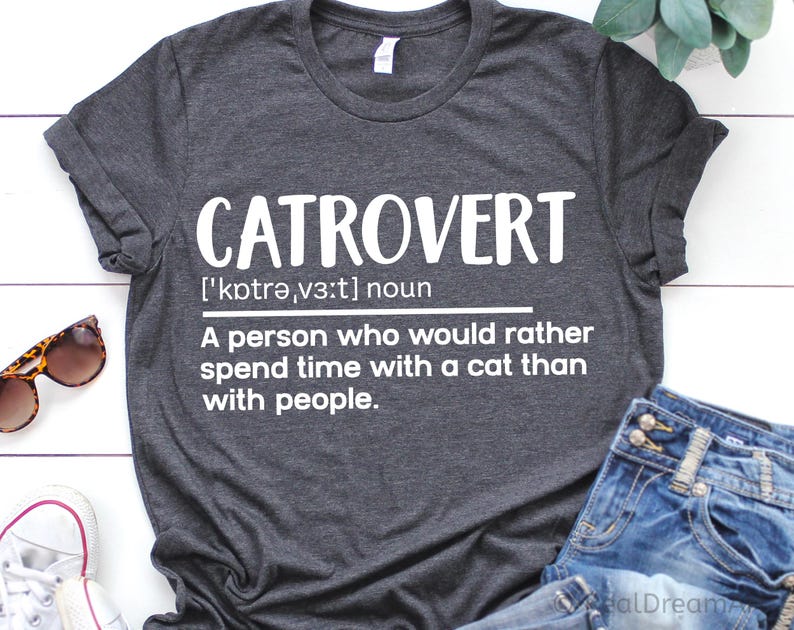 May include: Dark gray t-shirt with the word "CATROVERT" in large white letters. The definition below reads: "A person who would rather spend time with a cat than with people." Sunglasses and jeans are visible.