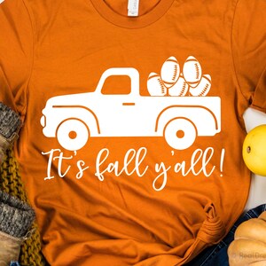 May include: Orange t-shirt with a white graphic of a pickup truck carrying footballs. The text "It's fall y'all!" is written below the truck.