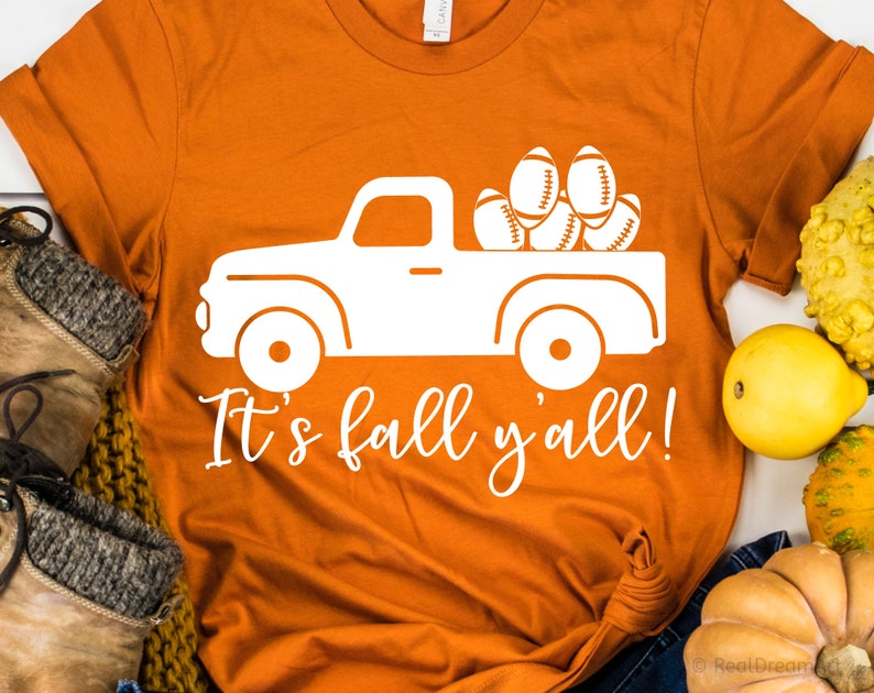 May include: Orange t-shirt with a white graphic of a pickup truck carrying footballs. The text "It's fall y'all!" is written below the truck.