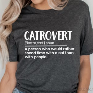 May include: Dark gray t-shirt with the word "CATROVERT" in large white letters. Below the word is the phonetic pronunciation and definition: "A person who would rather spend time with a cat than with people."