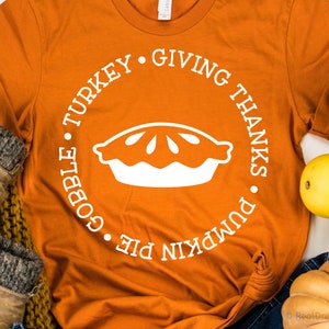 May include: Orange t-shirt with a white circle design featuring a pie, the words "pumpkin pie", "gobble", "turkey", "giving thanks" and a white outline.