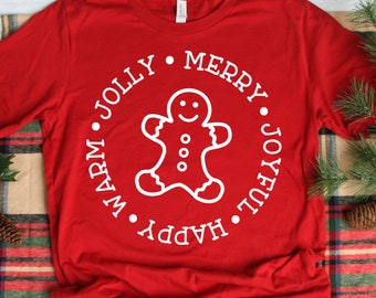 Op de afbeelding: Rood langarm T-shirt met een witte gingerbread man afbeelding en de tekst "Warm. Happy. Jolly. Merry. Joyful." rond de gingerbread man.