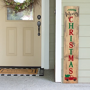 May include: A wooden sign with the words "Merry Christmas" in green and red letters. The sign also features a red truck with a Christmas tree in the back. The sign is leaning against a white door with a wreath on it.