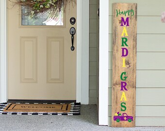 May include: A wooden sign with the words "Happy Mardi Gras" in purple, yellow, and green letters. A purple and gold fleur-de-lis is above a purple truck with a load of fleur-de-lis.