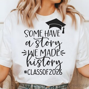Pu&ograve; includere: T-shirt bianco con testo nero e un'immagine di cappello di laurea. Il testo recita "SOME HAVE a STORY WE MADE history #CLASSOF2026". Una maglietta casual per le celebrazioni di laurea.