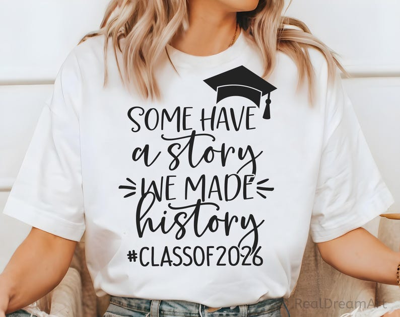 Pu&ograve; includere: T-shirt bianco con testo nero e un'immagine di cappello di laurea. Il testo recita "SOME HAVE a STORY WE MADE history #CLASSOF2026". Una maglietta casual per le celebrazioni di laurea.