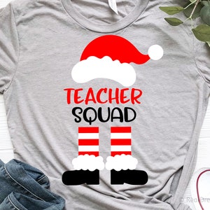 May include: A gray t-shirt with a white and red Santa hat and legs design. The text "Teacher Squad" is printed in black on the shirt.