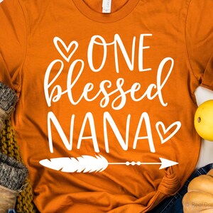 May include: Orange t-shirt with white text that reads "One Blessed Nana" with a heart on either side of the text. A white feather with an arrow is below the text.