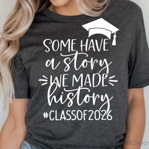 Pu&ograve; includere: T-shirt grigio scuro con testo bianco e un cappello di laurea. Il testo recita "Some have a story, we made history, #Classof2026". Una maglietta casual per le celebrazioni di laurea.