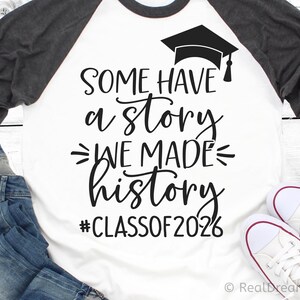 Pu&ograve; includere: Maglietta da baseball raglan bianca e grigia con la scritta "SOME HAVE a story WE MADE history #CLASSOF2026" e una grafica di cappello di laurea. La maglietta &egrave; abbinata a jeans blu e scarpe da ginnastica bianche.