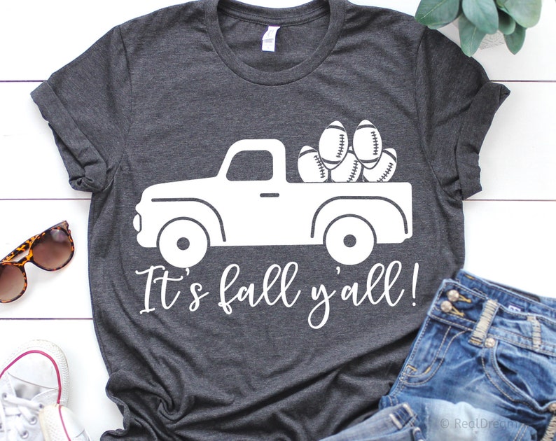 May include: A dark gray t-shirt with a white graphic of a pickup truck carrying footballs. The text "It's fall y'all!" is printed below the truck.