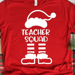 May include: A red long-sleeve shirt with a white graphic design. The design features a Santa hat, the words "TEACHER SQUAD", and elf legs with striped socks and shoes. The shirt is a festive holiday item.