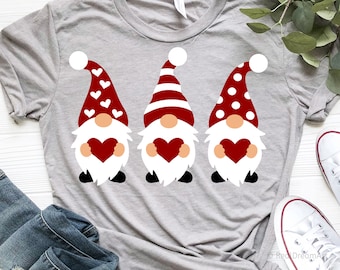 May include: Three gnomes wearing red, white, and red and white striped hats holding red hearts. The gnomes are on a gray t-shirt.