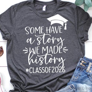 Pu&ograve; includere: T-shirt grigio scuro con testo bianco: "SOME HAVE a STORY WE MADE history #CLASSOF2026" e grafica del cappello di laurea. La maglietta &egrave; esposta con occhiali da sole e jeans.