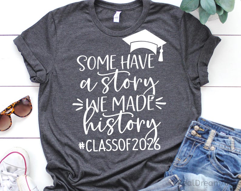 Pu&ograve; includere: T-shirt grigio scuro con testo bianco: "SOME HAVE a STORY WE MADE history #CLASSOF2026" e grafica del cappello di laurea. La maglietta &egrave; esposta con occhiali da sole e jeans.