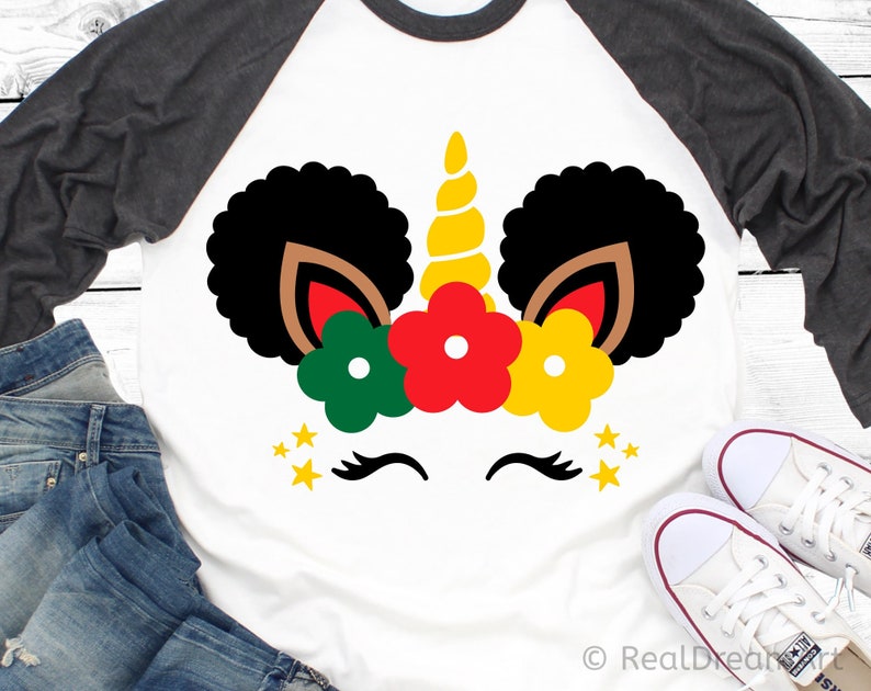 May include: A white t-shirt with a black and brown unicorn design. The unicorn has a gold horn and is wearing a crown of red, yellow, and green flowers. There are gold stars around the unicorn's face.