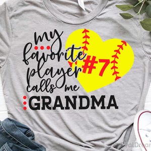 May include: A gray t-shirt with the text "my favorite player calls me GRANDMA" in black script. A yellow softball heart with red stitching and the number 7 is on the right side of the shirt.