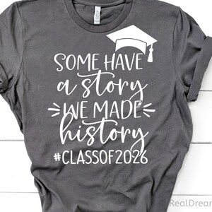 Pu&ograve; includere: T-shirt grigio scuro con testo bianco e grafica del cappello di laurea. Il testo recita "SOME HAVE a story WE MADE history #CLASSOF2026". La maglietta &egrave; piegata, mostrando le maniche arrotolate.