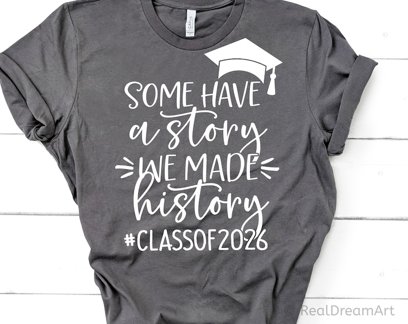 Pu&ograve; includere: T-shirt grigio scuro con testo bianco e grafica del cappello di laurea. Il testo recita "SOME HAVE a story WE MADE history #CLASSOF2026". La maglietta &egrave; piegata, mostrando le maniche arrotolate.