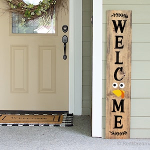 May include: A wooden welcome sign with a turkey face design. The sign is brown with black lettering and a black turkey face with yellow beak and orange wattle. The sign is leaning against a white door.