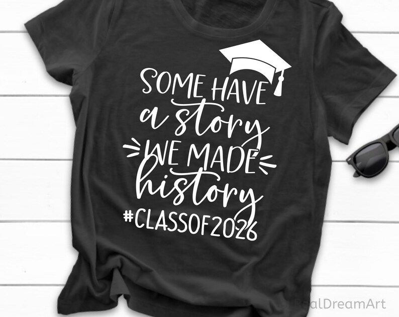 Pu&ograve; includere: T-shirt nero con testo bianco: "SOME HAVE a story WE MADE history #CLASSOF2026" e grafica di cappello di laurea. Un paio di occhiali da sole neri sono appoggiati sul lato della maglietta piegata.