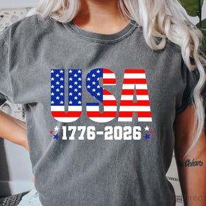 May include: A charcoal grey short-sleeve t-shirt featuring the text USA in a red, white, and blue American flag pattern. Below the text, the numbers 1776-2026 are printed in white, flanked by small red and blue stars on the left and right sides.