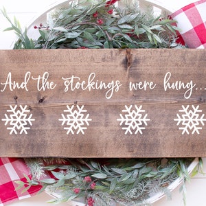 Puede incluir: Un letrero de madera con el texto "And the stockings were hung..." en letras blancas cursivas. Cuatro diseños de copos de nieve blancos se muestran en el letrero.