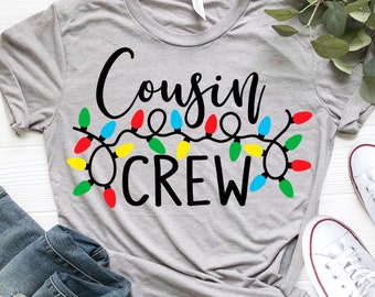 Puede incluir: Camiseta gris con el texto "Cousin Crew" en letras negras. El texto está rodeado de una cadena de luces navideñas de colores.