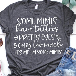 Puede incluir: Camiseta gris oscuro con texto blanco: "SOME MIMIS have tattoos, PRETTY EYES & cuss too much, IT'S ME, I'M SOME MIMIS." Gafas de sol y shorts vaqueros visibles. Una camiseta gráfica informal.