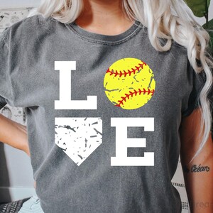 May include: A gray t-shirt with the word "LOVE" printed in white. The "O" is replaced with a yellow softball with red stitching. The "V" is replaced with a white silhouette of a baseball home plate.