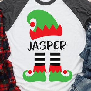 May include: White t-shirt with a green elf hat, red and white trim, black and white striped legs, and the name "Jasper" printed on the front.