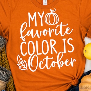 Puede incluir: Una camiseta naranja de manga corta con el texto en blanco MY favorite COLOR IS October, acompañado de un icono de calabaza en líneas blancas y un gráfico de hoja blanca. La camiseta se muestra junto a unas botas y pequeñas calabazas.