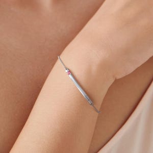 May include: A delicate silver bracelet featuring a rectangular bar with a small pink gemstone. The bracelet is worn on a wrist, showcasing its minimalist design and elegant appeal. The bracelet is a piece of jewelry.