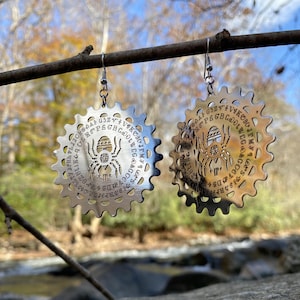 May include: A pair of silver earrings shaped like a circular wheel with a spider design and the letters of the alphabet around the edge. The earrings are hanging from a branch.