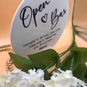 May include: White round sign with black text that reads "Open Bar" with a heart symbol. Below the text it reads "Tonight's Drinks Are Free, Tomorrow's Memories Are Priceless, Cheers!"