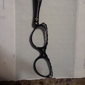 Puede incluir: Gafas de lectura negras con una montura y un mango decorativos. El mango y la montura están adornados con pequeños y brillantes pedrería. Las gafas tienen un pequeño lazo en la parte superior del mango.