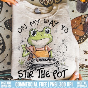 May include: A beige sweatshirt with a cartoon frog stirring a pot, with the text "ON MY WAY TO STIR THE POT". The frog is green and wearing orange overalls. The design includes small flowers and grass.