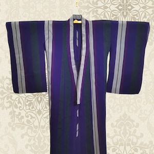 May include: A purple kimono with vertical stripes in shades of gray, green, and white. The garment is hanging on a gold hanger against a patterned background. The kimono has wide sleeves and a wrap-around design.