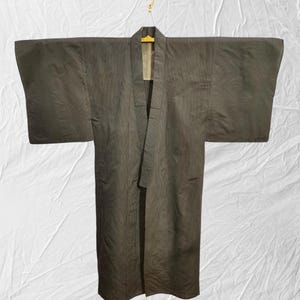 May include: A dark olive-green kimono hangs from a gold hanger against a white backdrop. The traditional Japanese garment features wide sleeves and a long, flowing design. The fabric appears to have subtle vertical stripes.