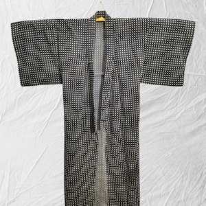May include: A black and white patterned kimono hangs on a gold hanger. The kimono has a repeating pattern of small white squares on a black background. The garment is open at the front and has wide sleeves.
