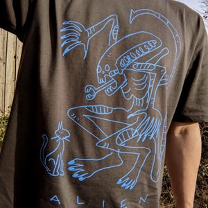 Peut inclure: T-shirt gris fonc&eacute; avec un dessin au trait bleu d'un extraterrestre et d'un chat. Le mot "ALIEN" est imprim&eacute; en bleu en bas. Le design est un graphisme artistique unique.