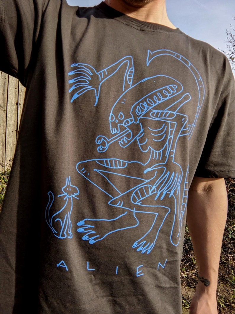 Peut inclure: T-shirt gris fonc&eacute; avec un dessin au trait bleu d'un extraterrestre et d'un chat. Le mot "ALIEN" est imprim&eacute; en bleu en bas. Le design est un graphisme artistique unique.
