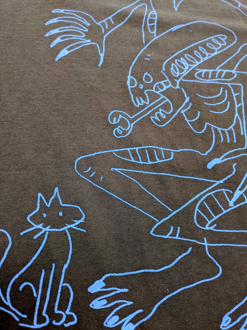 Peut inclure: T-shirt marron fonc&eacute; avec un dessin au trait bleu d'une cr&eacute;ature extraterrestre tenant un os et un chat. L'illustration est un design simple de type dessin anim&eacute;.