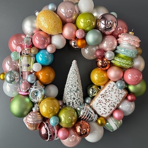 May include: A Christmas wreath made of colorful ornaments, including a gingerbread man, a candy cane, a lemon, a tree, and a macaron. The wreath is decorated with pink, green, blue, yellow, and white ornaments.