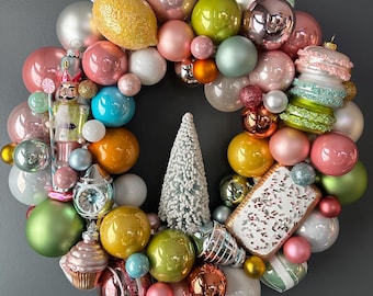 May include: A Christmas wreath made of colorful ornaments, including a gingerbread man, a candy cane, a lemon, a tree, and a macaron. The wreath is decorated with pink, green, blue, yellow, and white ornaments.