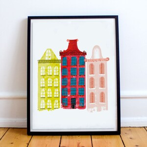 May include: A framed print of three colorful buildings with different architectural styles. The buildings are painted in shades of yellow, red, and pink, with blue windows. The print is a modern and abstract interpretation of a city scene.