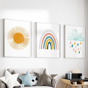Op de afbeelding: Een lichtgrijze kinderbank met vier kussens, twee grijs, een blauw en een wit met een zwart-wit pandabeerontwerp. De bank staat in een kamer met een houten bureau en stoel, een wit konijnenbeeldje en drie ingelijste prints aan de muur. De prints tonen een gele zon, een regenboog en een blauwe wolk met regendruppels.